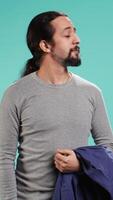 Vertical Man holding jacket in one hand while looking around, contemplating, creating a sense of curiosity. Person holding coat showing curiosity for surroundings, studio background, camera B video
