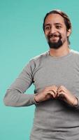 Vertical Portrait of joyful friendly man doing heart symbol shape gesture with hands. Smiling nurturing person showing love gesturing, isolated over blue studio background, camera B video