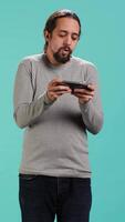 Vertical Focused man plays intense videogame on mobile phone, spamming attack buttons on touchscreen. Gamer participates in gaming competition, isolated over studio background, camera A video