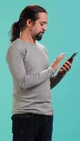 Vertical Portrait of smiling man scrolling on smartphone touchscreen, using mobile phone to check social media feed. Cheerful person browsing online websites, isolated over studio background, camera A video
