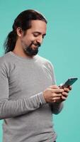 Vertical Happy man texting best friend using smartphone, reading messages. Relaxed person having fun while chatting with mate on mobile phone, isolated over blue studio background, camera B video