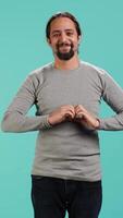 Vertical Portrait of joyful friendly man doing heart symbol shape gesture with hands. Smiling nurturing person showing love gesturing, isolated over blue studio background, camera A video