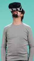Vertical Excited tech fan wearing virtual reality goggles, looking around. Arab man using high tech futuristic modern VR glasses, making tactile touching gesturing, studio background, camera B video