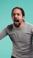 Vertical Portrait of irate man feeling outraged, yelling at friend, releasing pent up emotion, studio backdrop. Furious person full of rage screaming at mate isolated over blue studio background, camera B video