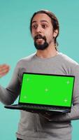Vertical Tech content creator doing influencer marketing using green screen laptop, isolated over studio background. Arab man holding empty copy space mockup notebook, doing review, camera B video