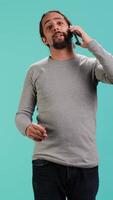 Vertical Joyous man catching up with friends in phone call using cellphone, studio background. Portrait of radiant person having conversation with mates during telephone call, camera A video