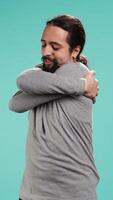 Vertical Portrait of man standing in studio, hugging himself in gesture of comfort and self love, isolated over studio background. Cheerful person doing self acceptance, embracing himself, camera B video