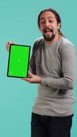 Vertical Happy man holding green screen tablet, doing recommendation. Cheerful person presenting mockup device, showing positive feedback, isolated over studio background, camera A video