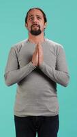Vertical Portrait of man putting hands together in begging gesture, making wish, hoping for good news isolated over studio background. Person waiting for results, praying for positive outcome, camera A video