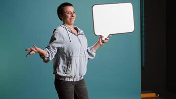 Female model with a groovy style holding a speech bubble in studio, creating an ad commercial with an empty billboard icon. Cool edgy woman presenting a sign for advertisement. Camera A. video