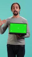 Vertical Tech content creator doing influencer marketing using green screen laptop, isolated over studio background. Arab man holding empty copy space mockup notebook, doing review, camera A video