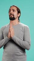 Vertical Portrait of man putting hands together in begging gesture, making wish, hoping for good news isolated over studio background. Person waiting for results, praying for positive outcome, camera B video