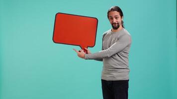 Smiling man holding red speech bubble sign of empty copy space for message. Joyous person presenting thought bubble cardboard used as promotion concept, studio background, camera A video