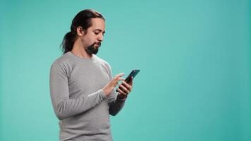 Portrait of smiling man scrolling on smartphone touchscreen, using mobile phone to check social media feed. Cheerful person browsing online websites, isolated over studio background, camera B video