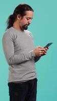 Vertical Happy man texting best friend using smartphone, reading messages. Relaxed person having fun while chatting with mate on mobile phone, isolated over blue studio background, camera A video