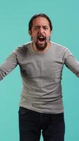 Vertical Portrait of irate man feeling outraged, yelling at friend, releasing pent up emotion, studio backdrop. Furious person full of rage screaming at mate isolated over blue studio background, camera A video