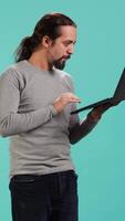 Vertical Upset man frowns while looking at laptop, dissatisfied with company profits, studio backdrop. Irked worker flabbergasted by low business income revenue, doing financial analysis on notebook, camera A video
