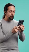 Vertical Man holding mobile phone, browsing betting website, celebrating after winning money, isolated over studio background. Cheerful person feeling happy while scrolling on smartphone, camera B video
