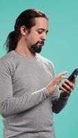 Vertical Portrait of smiling man scrolling on smartphone touchscreen, using mobile phone to check social media feed. Cheerful person browsing online websites, isolated over studio background, camera B video