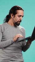 Vertical Upset man frowns while looking at laptop, dissatisfied with company profits, studio backdrop. Irked worker flabbergasted by low business income revenue, doing financial analysis on notebook, camera B video