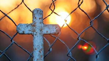 A rusty cross on a chain link fence with the sun setting behind it video