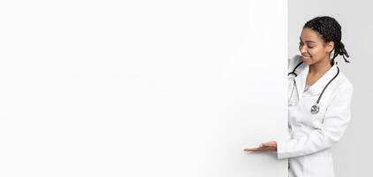 Free place for your ad. Young black female doctor looking and pointing at big blank advertising board with open palm, empty space for text, panorama photo