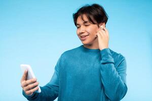 Asian young guy stands against a blue backdrop, smiling as he uses his smartphone and wears wireless earbuds. He appears to be enjoying music or possibly a call. photo