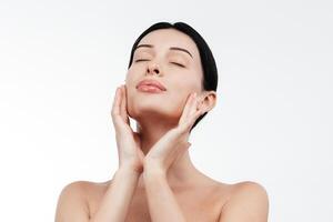 A young woman in a studio against a white background gently touches her flawless skin. Her expression conveys joy and harmony, highlighting her allure and well-groomed appearance photo