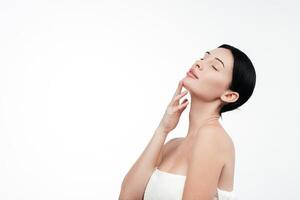 A young woman in a studio against a white background gently touches her flawless skin. Her expression conveys joy and harmony, highlighting her allure and well-groomed appearance photo