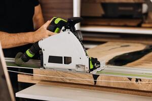 Unrecognizable man working with professional, electric circular saw, making a precise cut along the guide on a wooden surface. Tool is connected to a dust extraction system. Woodworking workshop. photo