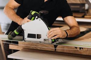 Unrecognizable man working with professional, electric circular saw, making a precise cut along the guide on a wooden surface. Tool is connected to a dust extraction system. Woodworking workshop. photo