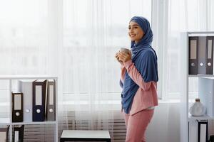 Break in work. Arabic female manager in hijab standing with coffee mug in office, relaxing against window at workplace, free space photo