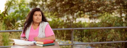 A young student sits at a table outside, surrounded by greenery, focused on her studies. She has books and notes open, enjoying the sunshine while working diligently on her assignments, panorama photo