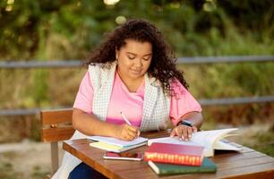 A student engages in studying at a wooden table outside, surrounded by nature. She writes in her notebook while several books lie open, showcasing her dedication to learning. photo