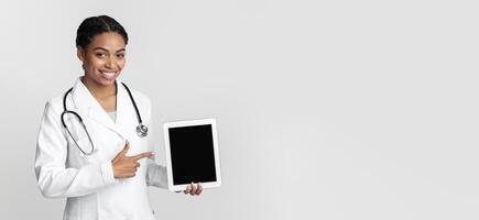 Medical App. Afro Female Doctor In medical Coat Pointing At Digital Tablet With Blank Screen In Her Hands, Standing Over Light Background, Mockup photo