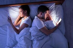 Infidelity. Cheating Spouses Using Smartphones Texting With Lovers Lying Back-To-Back In Bed At Home At Night. Top View, Low-Light photo
