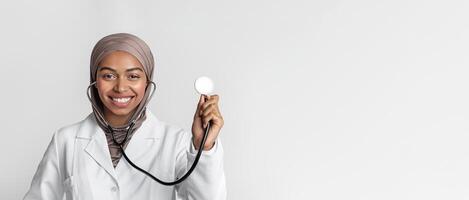 Portraif of young black muslim female doctor in hijab with stethoscope, posing over light background and smiling at camera, free space photo