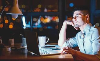 Boring Job. Overworked Business Guy Sitting At Laptop Uninterested In Work In Modern Office At Night. Low Light photo