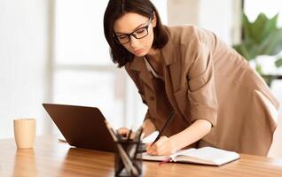Self employed female entrepreneur is busy writing in her notebook, standing at desk with large window in the background photo