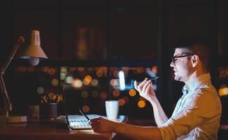 Business Lifestyle. Man Using Smartphone Voice Assistant Working On Laptop In Modern Office At Night. Copy Space photo