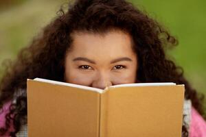 A lady with curly hair smiles while holding a book in front of her face, sitting on grass. The sun shines brightly, creating a cheerful atmosphere as she enjoys her reading time. photo