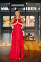 Portrait of a smiling girl in a red dress stands in the interior with her hands clasped in front of her photo