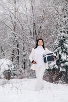 A stylish woman with a white suit with a New Year's gift in her hands in a winter forest. A girl in nature in a snowy forest with a gift box photo