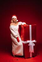 portrait of a smiling girl in a white dress and Santa hat with a huge Christmas gift on a red background photo