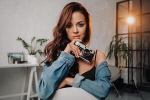 a brunette with long hair poses in a studio sitting on a chair with a camera in her hands photo