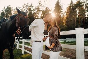 a pregnant girl in a hat and a man in white clothes stand next to horses at a White fence.Stylish family waiting for a child strolling in nature photo