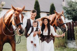 A family in white clothes with their son stand near two beautiful horses in nature. A stylish couple with a child are photographed with horses photo