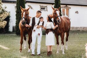 A family in white clothes with their son stand near two beautiful horses in nature. A stylish couple with a child are photographed with horses photo