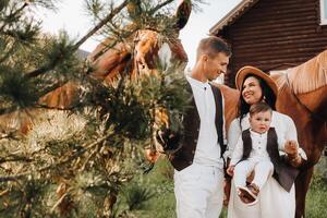 A family in white clothes with their son stand near two beautiful horses in nature. A stylish couple with a child are photographed with horses photo