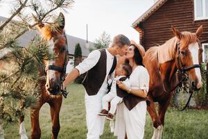 A family in white clothes with their son stand near two beautiful horses in nature. A stylish couple with a child are photographed with horses photo